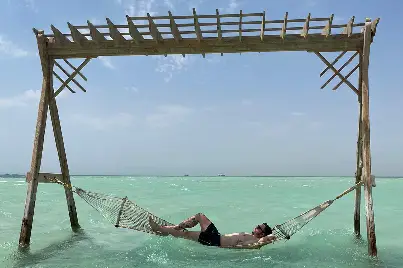 A tourist relaxing on a luxury wooden beach bed over the crystal-clear turquoise water at Orange Bay Island Hurghada.